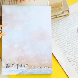 May include: A rectangular notepad with a winter scene featuring penguins sliding on ice. The background has a soft, watercolor effect with a light blue and pink color palette. The notepad is surrounded by vintage books and flowers.