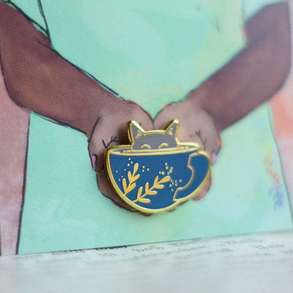 May include: A gold-tone enamel pin featuring a cat curled up inside a blue teacup. The cat has a white face and a black nose. The teacup has a gold rim and a gold handle. The teacup is decorated with gold leaves and dots.