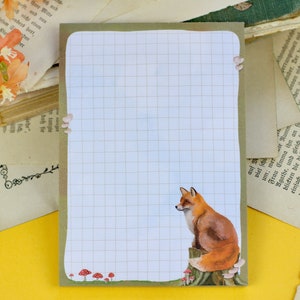 May include: A white gridded notepad with a brown border. The notepad features a fox illustration in the bottom right corner, sitting on a tree stump with mushrooms around it.
