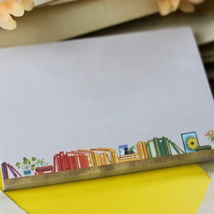 May include: A white notepad with a brown border and a colourful illustration of a bookshelf with books and plants at the bottom.