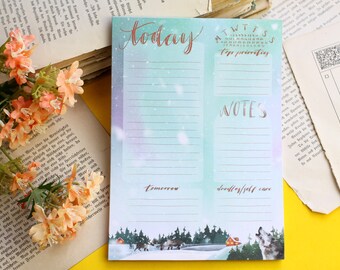 May include: A blue and white notepad with a wintery scene at the bottom. The notepad is divided into two sections, one labeled "today" and the other labeled "notes". The "today" section has a list of days of the week and a space for "top priorities". The "notes" section has a space for "tomorrow" and "doodles/self care".