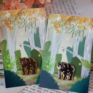 May include: Two enamel pins featuring a bear design, displayed on cards with a forest illustration and the text "Tiny Little Forest". The pins are gold and black with gold outlines. The background includes old books and floral accents.