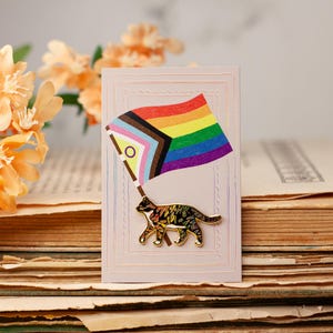 Peut inclure: Épinglette de chat tenant un drapeau Progress Pride, sur fond de vieux livres et de fleurs orange. L'épinglette présente un drapeau coloré et une silhouette de chat détaillée. L'esthétique générale est vintage et artistique.
