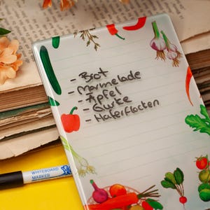 May include: A clear, rectangular whiteboard with a colourful vegetable design and a handwritten grocery list. A black marker and a stack of old books with flowers are in the background.