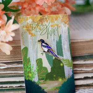 May include: An enamel pin with a magpie design, set against a forest scene with the words "Tiny Little Forest". The magpie has blue and white details. The background includes a stack of old books and orange flowers. The pin is a decorative accessory.