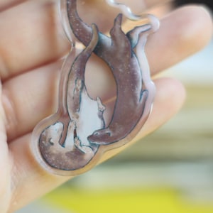 Könnte beinhalten: Ein klarer Acryl-Schlüsselanhänger mit einer Illustration von zwei sich umarmenden Ottern. Die Otter sind in Braun- und Weißtönen dargestellt und befinden sich in einer spielerischen Pose. Der Schlüsselanhänger hat ein glänzendes Finish.
