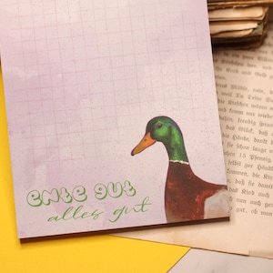 Könnte beinhalten: Ein Notizblock mit Aquarellhintergrund und Rastermuster. Der Notizblock zeigt eine farbenfrohe Illustration einer Ente mit grünem Kopf, braunem Körper und orangem Schnabel. Der Text "ente gut alles gut" ist grün geschrieben.