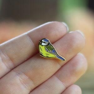 Könnte beinhalten: Eine goldfarbene Emaille-Anstecknadel mit einem Blaumeisen-Vogel mit gelber Brust, blauen Flügeln und schwarzem Kopf. Der Vogel sitzt auf einem Ast.
