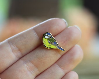May include: A gold-colored enamel pin featuring a blue tit bird with a yellow breast, blue wings, and a black head. The bird is perched on a branch.