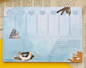 May include: A blue weekly planner with cat illustrations. The planner has a grid layout with days of the week labeled M, T, W, T, F, S, and S. Each day has a space for writing. The planner has a light blue background with a watercolor effect. There are cats in various poses throughout the planner.