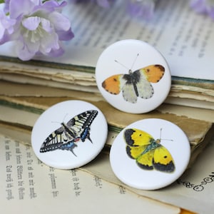 Könnte beinhalten: Drei weiße runde Knöpfe mit Schmetterlingsillustrationen. Einer zeigt einen gelb-schwarzen Schmetterling, ein anderer einen orange-weißen Schmetterling und der dritte einen schwarz-gelb-blauen Schmetterling. Die Knöpfe liegen auf einem Stapel alter Buchseiten.