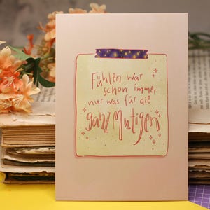 Peut inclure: Une carte rose avec le texte allemand "Fühlen war schon immer nur was für die ganz Mutigen" en script rouge. La carte est scotchée avec du ruban adhésif washi violet et se trouve devant une pile de vieux livres et de fleurs orange.