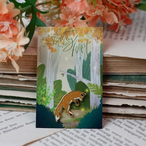 Könnte beinhalten: Ein goldfarbener Fuchs-Emaille-Pin auf einer Karte mit dem Text "Tiny Little Forest". Die Karte zeigt eine Waldszene mit Bäumen und Blättern. Der Pin befindet sich vor einem Hintergrund aus alten Büchern und pfirsichfarbenen Blumen.