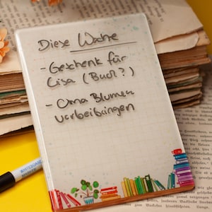 Könnte beinhalten: Ein weißer Notizblock mit Rastermuster und handschriftlichem deutschen Text, darunter "Diese Woche". Der Notizblock ist mit einem Stapel bunter Bücher und einer Pflanzenillustration verziert. Ein schwarzer Marker und ein Stapel alter Bücher sind ebenfalls sichtbar.