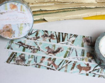 May include: A roll of washi tape with a blue and white background featuring a repeating pattern of squirrels in a snowy forest. The tape is partially unrolled and the text "Squirrels & Snowflakes" is visible on the roll.