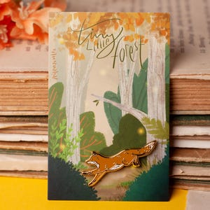 Könnte beinhalten: Eine goldfarbene Emaille-Anstecknadel in Form eines rennenden Fuchses, befestigt auf einer rechteckigen Karte mit einer Waldillustration, weißen Bäumen, grünem Blattwerk und dem Schriftzug Tiny Little Forest in schwarzer Schreibschrift.
