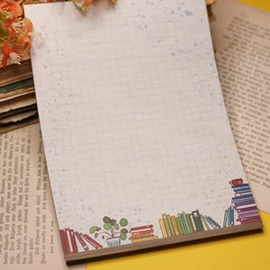 Peut inclure: Un bloc-notes blanc avec un motif quadrillé et une bordure de livres colorés et une plante en pot. Le bloc-notes a un dos brun clair.