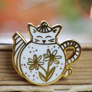 Può includere: Spilla in smalto dorata con una teiera a forma di gatto bianca con un motivo floreale. La teiera ha un piccolo cappello sulla testa e un manico a righe.