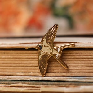 Könnte beinhalten: Goldfarbene Emaille-Anstecknadel mit einem stilisierten Schwalbenmotiv. Der Vogel ist im Flug dargestellt, mit weit ausgebreiteten Flügeln. Die Nadel hat eine weiße und goldene Farbgebung.