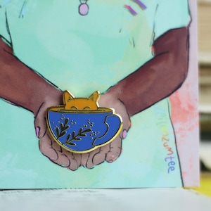 Könnte beinhalten: Eine goldene Emaille-Anstecknadel mit einer Katze, die aus einer blauen Teetasse mit goldener Verzierung und grünen Blumenmotiven schaut. Die Anstecknadel wird in den Händen einer Person im Hintergrund gehalten.