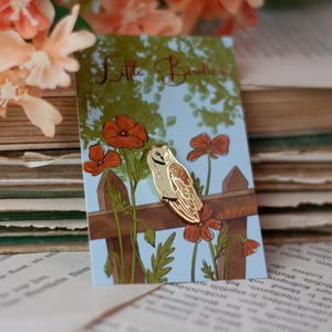 Può includere: Spilla a forma di gufo color oro su una carta con sfondo blu, una staccionata in legno e fiori arancioni. La carta recita "Little Birdies". La spilla è ambientata su uno sfondo di vecchi libri e fiori color pesca.