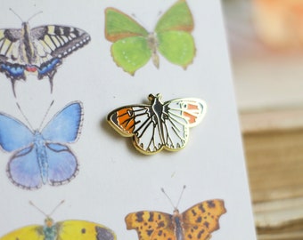 May include: Gold-tone enamel pin featuring a white butterfly with orange and black accents. The butterfly is set against a white background with illustrations of other butterflies.