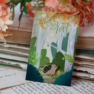 May include: A gold-colored hedgehog enamel pin with black and white details, set against a forest-themed card. The card features a forest illustration with the text "Tiny Little Forest". The pin is displayed on a stack of vintage books.