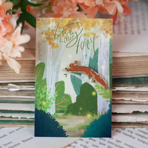May include: A pin featuring a red squirrel on a branch, set against a forest scene. The pin is on a card with the words "Tiny Little Forest" and a forest illustration. The background includes old books and peach-colored flowers.