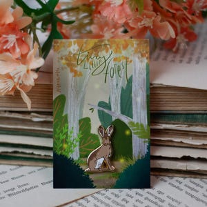 Könnte beinhalten: Ein Emaille-Pin mit einem braunen Hasen in einer Waldszene. Der Pin hat goldene Akzente und ist vor einem Hintergrund aus Bäumen und Blättern platziert. Auf der Karte steht "Tiny Little Forest".