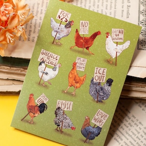 May include: A green greeting card featuring illustrations of chickens holding signs with various messages. The card is placed on a stack of old books with orange flowers in the background. The messages on the signs include "FCK NZS", "No Kings", and "Silence is Violence".