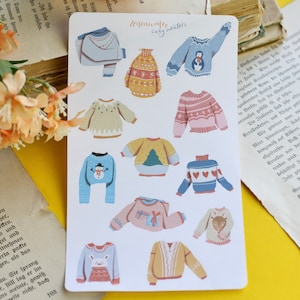 May include: A sticker sheet with illustrations of cosy sweaters in various colours and designs. The sweaters include patterns with hearts, a snowman, a Christmas tree, and a reindeer. The text "Momente cozy sweaters" is at the top.