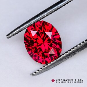 Op de afbeelding: Een gefacetteerde, ovaalvormige, dieprode edelsteen wordt vastgehouden door een zilveren pincet. De facetten van de edelsteen vangen het licht op, waardoor een schitterende glans ontstaat. De tekst "JEFF DAVIES & SON GEMSTONES & FACETING" is inbegrepen.