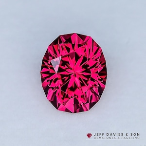 Puede incluir: Una gema facetada, de corte ovalado, con un tono rosa intenso y vibrante. La estructura interna de la gema refleja la luz, creando un efecto de estallido estelar. La imagen incluye el texto "JEFF DAVIES & SON GEMSTONES & FACETING".