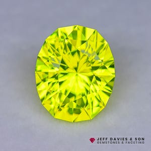May include: A faceted, oval-cut gemstone with a vibrant, neon-yellow hue. The gem's surface is a complex arrangement of triangular facets, reflecting light. The gemstone is set against a neutral gray background. The logo "JEFF DAVIES & SON GEMSTONES & FACETING" is in the bottom right corner.