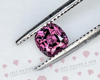 Può includere: Una pietra preziosa di colore rosa violaceo con taglio a cuscino, tenuta da una pinzetta metallica color argento. La pietra presenta una superficie sfaccettata che riflette la luce. Il testo JEFF DAVIES & SON GEMSTONES & FACETING è visibile in basso.