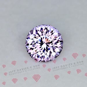 Pode incluir: Uma pedra preciosa redonda e facetada de cor roxo claro com um corte complexo e simétrico. A pedra exibe reflexos de luz geométricos e nítidos na sua superfície. O texto JEFF DAVIES & SON GEMSTONES & FACETING aparece abaixo.