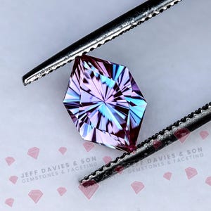 Puede incluir: Una gema facetada con un diseño geométrico complejo, sujeta con unas pinzas. La gema presenta una mezcla de tonos morados, azules y rosas, con un patrón de estrella que emana del centro. Las pinzas son de color plateado.