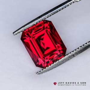 Può includere: Una gemma rossa vibrante, taglio rettangolare, è tenuta da una pinzetta d'argento. Le sfaccettature della gemma riflettono la luce, mostrando la sua chiarezza e brillantezza. L'immagine include il testo "JEFF DAVIES & SON GEMSTONES & FACETING".