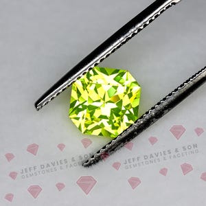 May include: A faceted, cushion-cut, vibrant lime-green gemstone held by metal tweezers. The background features a repeating pattern of small pink diamond icons and the text JEFF DAVIES & SON GEMSTONES & FACETING in gray lettering.