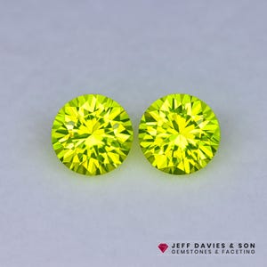 May include: Two round, faceted, bright yellow-green gemstones. The gems are set against a light gray background. The gemstones are likely cut to maximize light reflection and sparkle. The logo "JEFF DAVIES & SON GEMSTONES & FACETING" is in the bottom right corner.