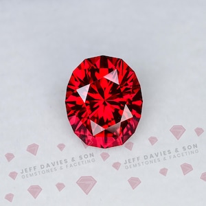 Puede incluir: Una gema roja vibrante de corte ovalado con una superficie compleja de múltiples facetas que refleja la luz en un patrón de estrella. La piedra se muestra sobre un fondo claro con el texto JEFF DAVIES & SON GEMSTONES & FACETING.