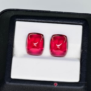 May include: Two cushion-cut, vibrant red gemstones displayed in a black jewelry box with a white interior. The gems have a high luster and are set side by side, showcasing their rich color and clarity. The gemstones are a deep, saturated red.