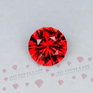 May include: A round, faceted, vibrant red gemstone with a brilliant cut, displaying sharp geometric light reflections. The text JEFF DAVIES & SON GEMSTONES & FACETING is printed below the stone, accompanied by small diamond-shaped icons.