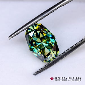 Peut inclure: Une pierre précieuse à facettes aux teintes vertes, jaunes et bleues, maintenue par des pinces métalliques. La taille de la gemme révèle de multiples facettes, créant un effet scintillant. Le logo "JEFF DAVIES & SON GEMSTONES & FACETING" est visible.