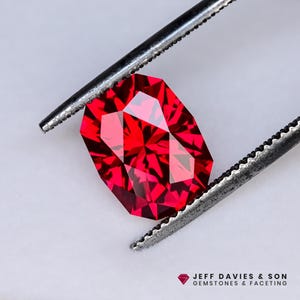 May include: A faceted, rectangular-cut, vibrant red gemstone is held by tweezers. The gem has multiple facets that reflect light, creating a sparkling effect. The image includes the text "JEFF DAVIES & SON GEMSTONES & FACETING".