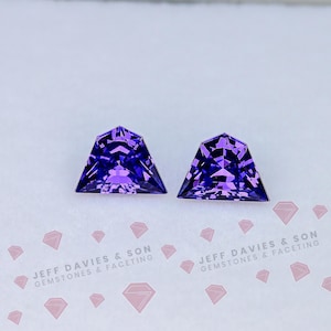 May include: Two triangular-shaped, faceted gemstones in a deep purple colour. The gems are cut with multiple facets, creating a sparkling effect. The gemstones are displayed on a white surface with the text "JEFF DAVIES & SON GEMSTONES & FACETING".