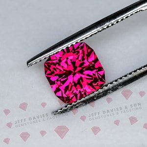 Könnte beinhalten: Ein leuchtender, fuchsiafarbener Edelstein im Kissenschliff, gehalten von einer silberfarbenen Metallpinzette. Der Stein weist komplexe, symmetrische Facetten auf, die das Licht reflektieren. Der Text JEFF DAVIES & SON GEMSTONES & FACETING ist unten sichtbar.