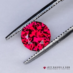 May include: A faceted, round, bright red gemstone held by a pair of metal tweezers. The gem has multiple facets that reflect light, creating a sparkling effect. The image includes the text "JEFF DAVIES & SON GEMSTONES & FACETING".