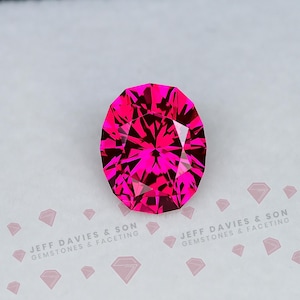 Puede incluir: Una gema de corte ovalado en un color magenta-rosa intenso y saturado, con facetas simétricas complejas que reflejan la luz. La piedra está colocada sobre un fondo blanco que presenta el texto JEFF DAVIES & SON GEMSTONES & FACETING.