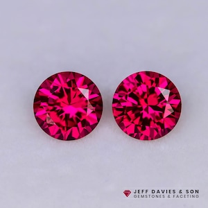 Puede incluir: Dos piedras preciosas redondas de color rojo vibrante, con talla brillante y facetas complejas que reflejan la luz. Las piedras están colocadas una al lado de la otra. El texto JEFF DAVIES & SON GEMSTONES & FACETING aparece abajo.